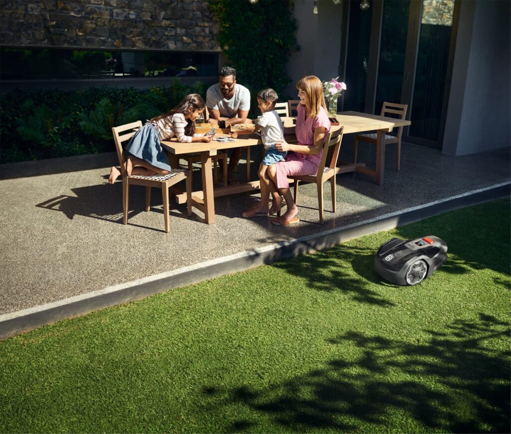 Familie sitzt auf Terrasse während Rasenroboter automatisch den Garten mäht